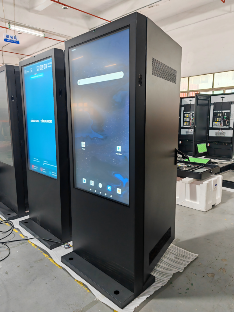 IP65 rated 43-inch outdoor digital signage kiosk undergoing pre-shipment quality control testing in manufacturing facility, featuring weatherproof enclosure and built-in cooling system