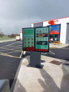 Outdoor double-sided digital menu board at fast food drive-thru with weatherproof touchscreen display showing menu items and pricing