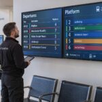 Ein Techniker prüft farbige Linienanzeigen und Abfahrtsinformationen auf zwei wandmontierten gewerblichen Displays.