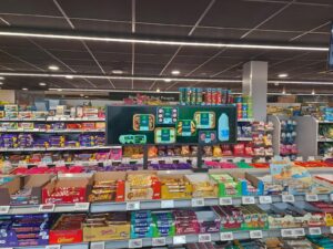 Stretch bar LCD display mounted on supermarket shelving showing fresh meal products and prepared food advertising above candy and snack aisles