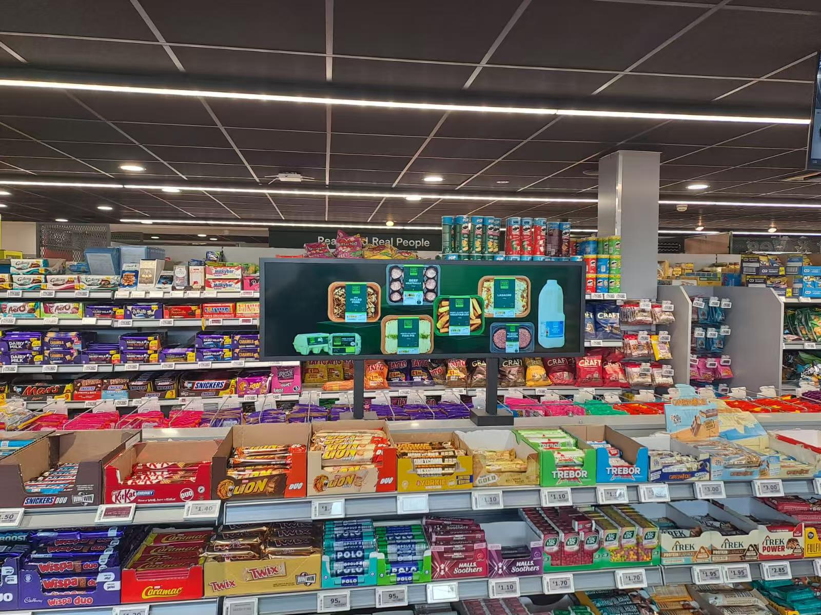 Stretch bar LCD display mounted on supermarket shelving showing fresh meal products and prepared food advertising above candy and snack aisles