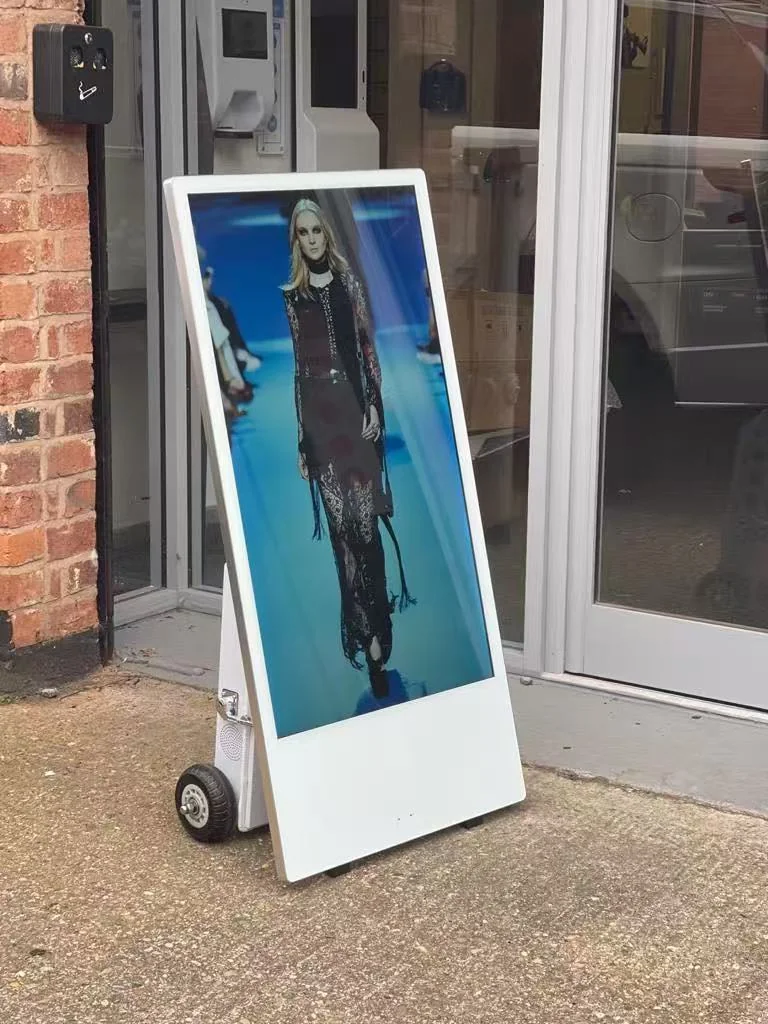 Weißes Display auf Rollen vor einem verglasten Gebäudeeingang, mit einer Modeaufnahme eines Models im Hochformat.