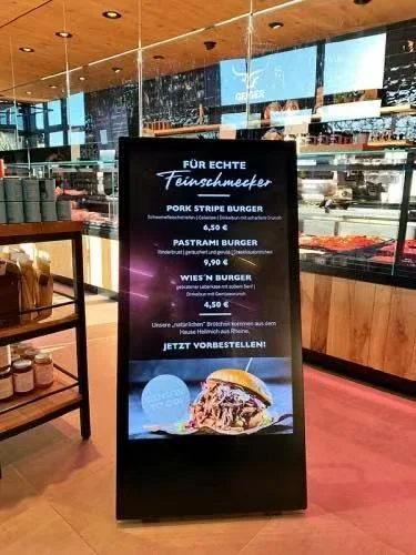 Schwarzes Display im Hochformat nahe einer Restauranttheke, mit einer Burgerkarte, Preisen und Speisenbildern.