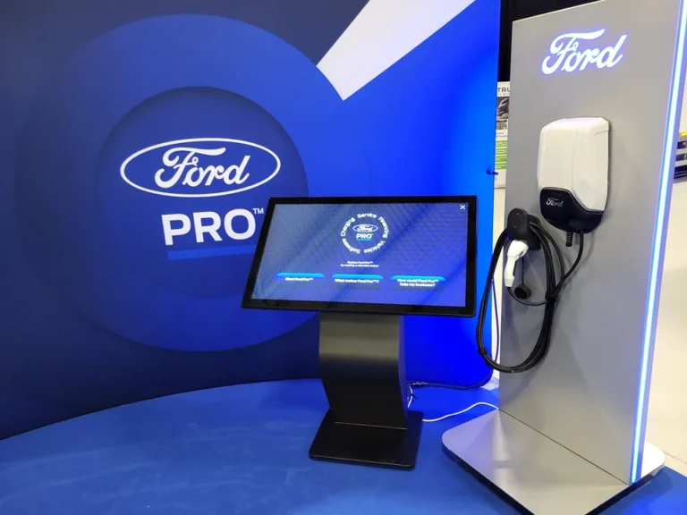 Terminal mit geneigtem Touchscreen am Ford Pro Messestand, mit Marken- und Serviceinformationen neben einer Fahrzeugladestation.