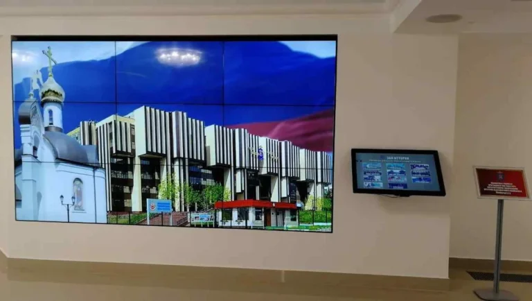 LCD-Videowand aus mehreren Panels in einem Firmen-Showroom, mit Gebäudeaufnahmen und Werbemotiven sowie einem kleineren Bildschirm daneben.