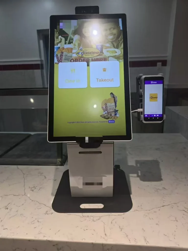 Bestellterminal auf einer Restauranttheke mit Auswahl für Verzehr vor Ort oder Mitnahme, Zahlungsterminal rechts und Belegdrucker darunter.