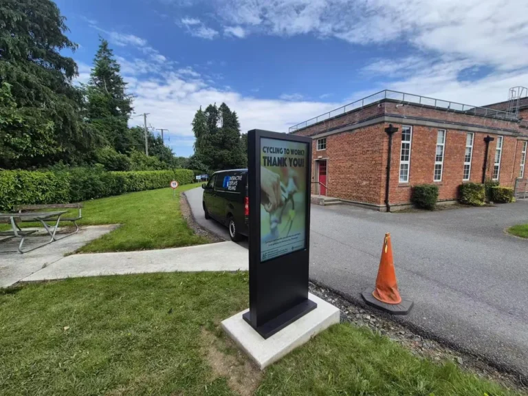 Black outdoor digital totem on a fixed base beside a building and lawn, displaying a cycle-to-work message.