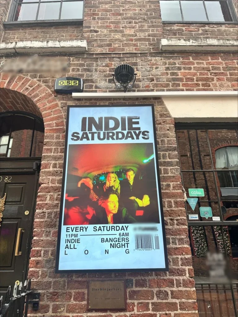 Portrait outdoor LCD display on a brick wall beside a venue entrance, showing a Saturday music event poster.