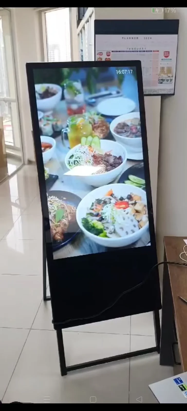 Black floor-standing display near restaurant doors and windows, showing food images, with a support stand and a connected power cable.