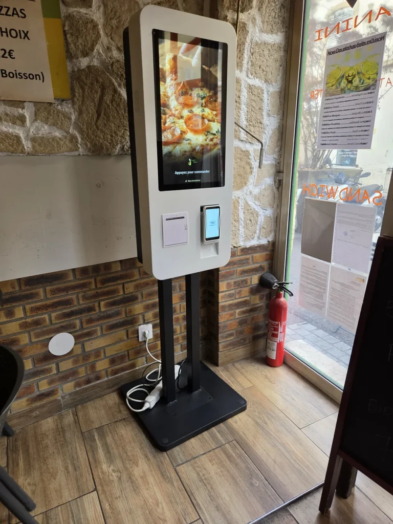 White floor-standing ordering kiosk near a restaurant entrance, with an ordering interface, payment terminal and receipt outlet.