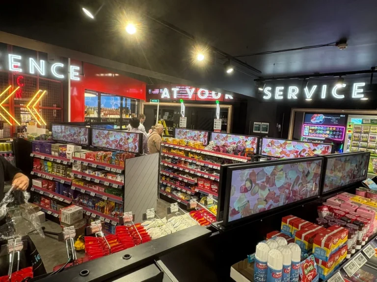 Horizontale LCD-Displays im Streifenformat über Supermarktregalen nahe den Kassen, mit Werbung für Süßwaren.
