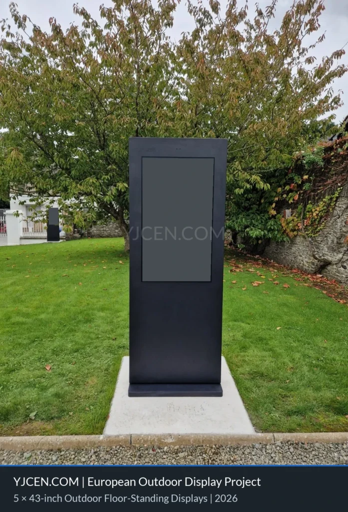 Black outdoor floor-standing display on a concrete base beside a lawn and gravel path, with screen content concealed for privacy.