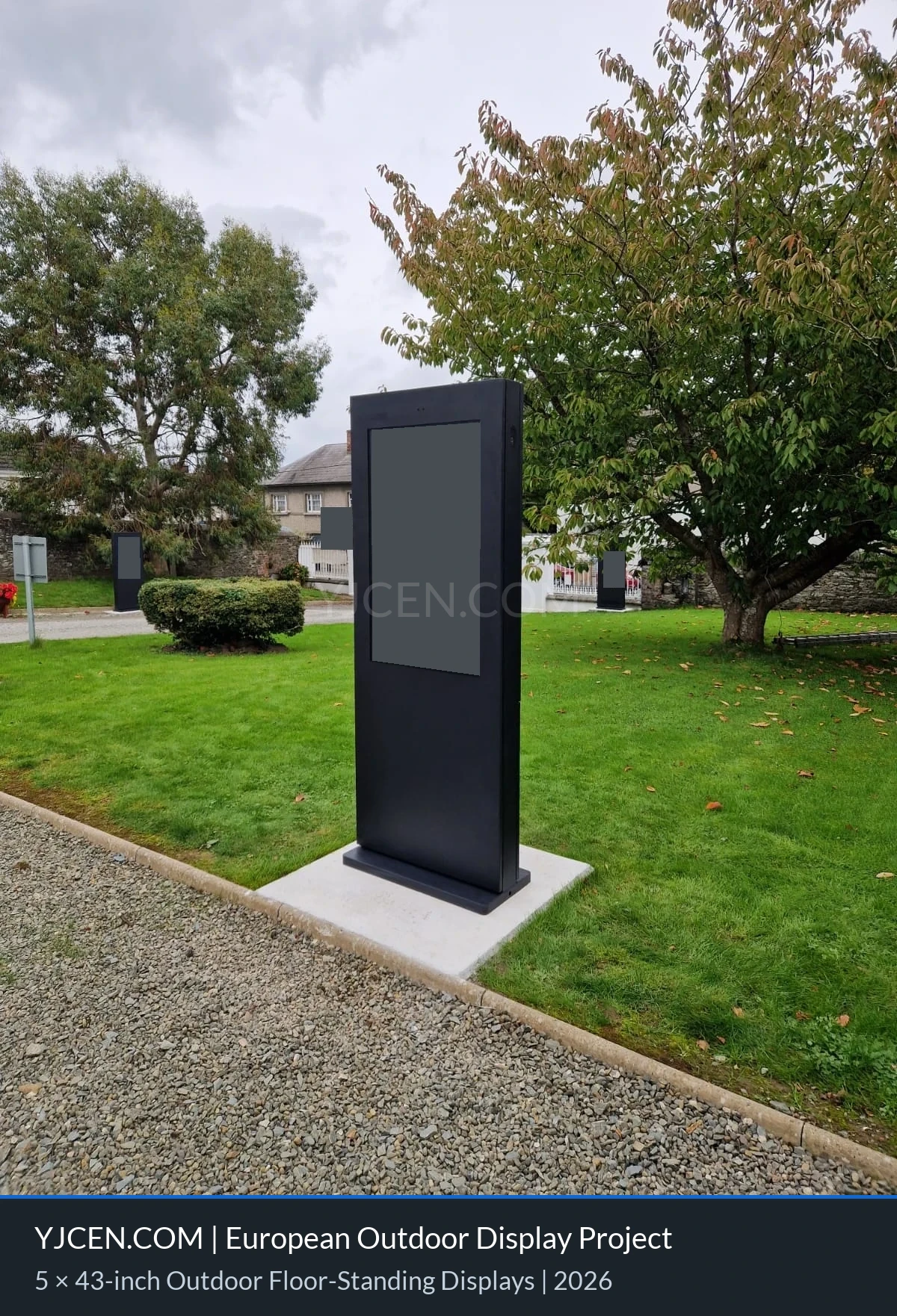 Vista em ângulo de um totem digital externo ao lado de um gramado, com o conteúdo das telas e as placas ao fundo ocultados por confidencialidade.