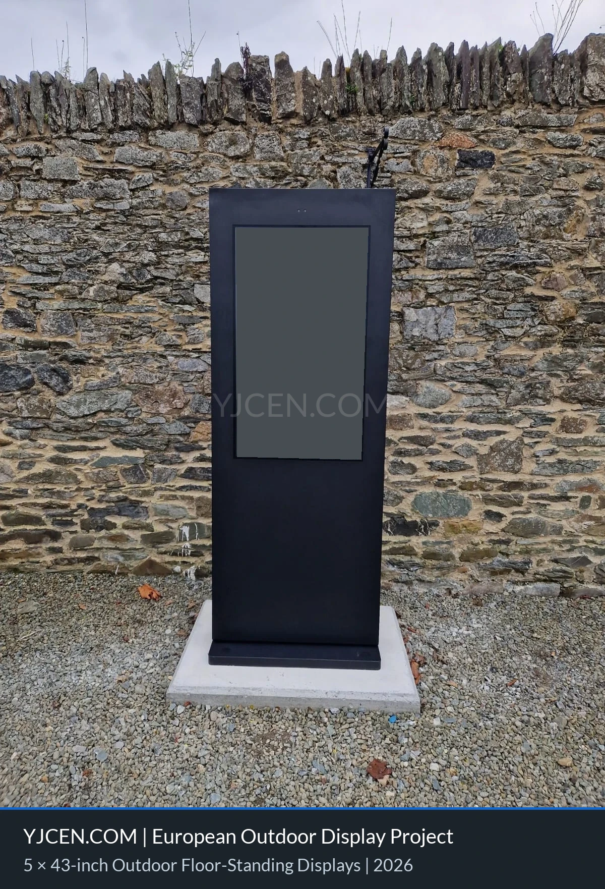 Vista frontal aproximada de um totem digital externo ao lado de um muro de pedra, com o conteúdo da tela ocultado por confidencialidade.