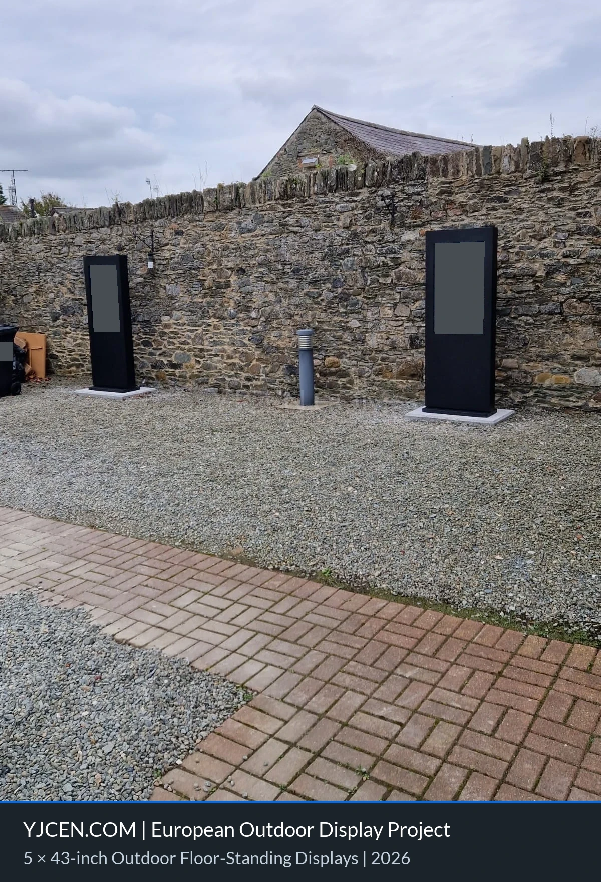 Dois totens digitais externos ao longo de um muro de pedra, com o conteúdo das telas ocultado por confidencialidade.