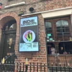 75-Zoll-IP66-Outdoor-Display im Hochformat neben dem Eingang eines europäischen Musikclubs mit Veranstaltungsmotiv.