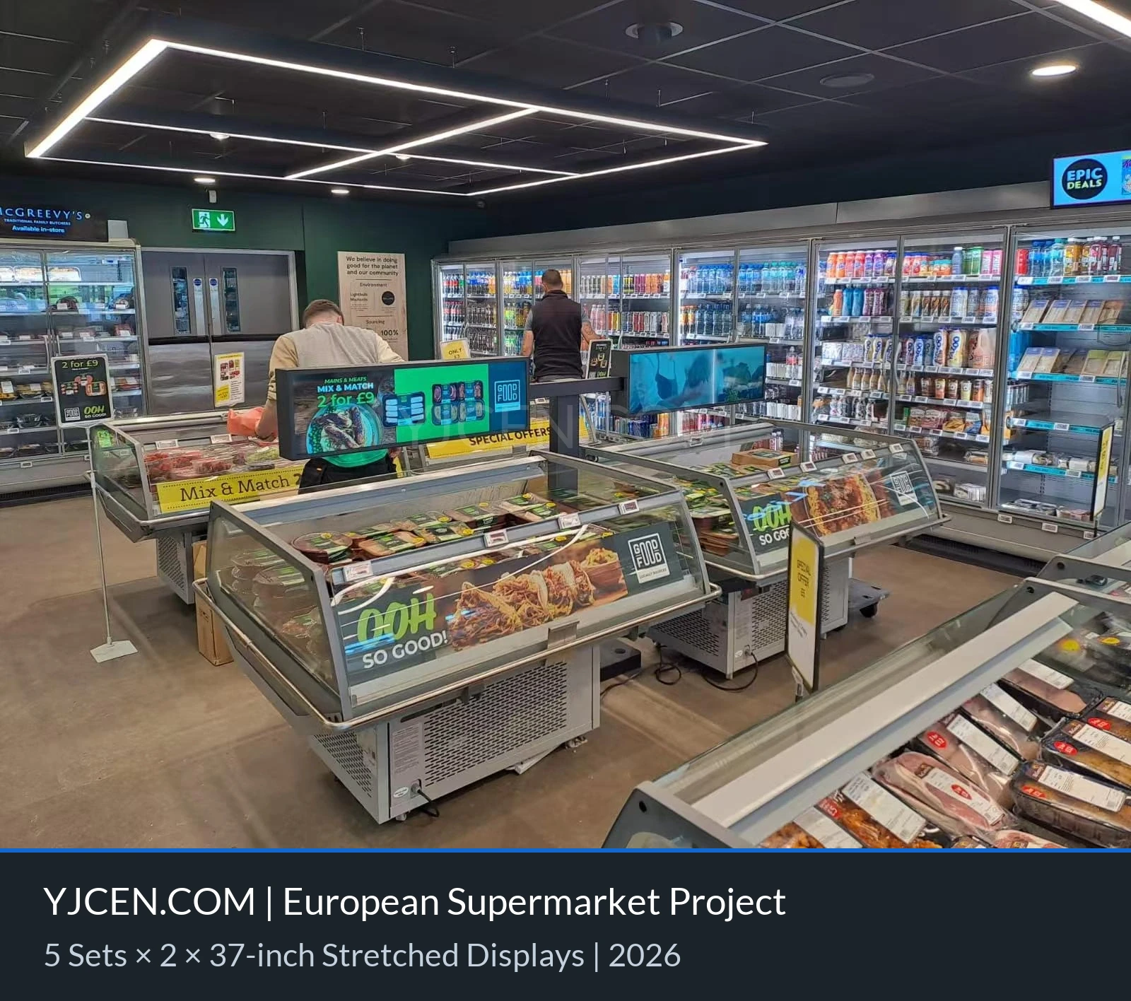 Vista frontal de dois displays em formato de barra de 37 polegadas exibindo promoções e conteúdo de vídeo acima dos expositores refrigerados de um supermercado.