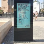 كشك توجيه رقمي خارجي قائم بذاته مقاس 55 بوصة في ساحة عامة بمراكش، يعرض شاشة ترحيب بمعلومات سياحية متعددة اللغات مع مئذنة الكتبية في الخلفية.