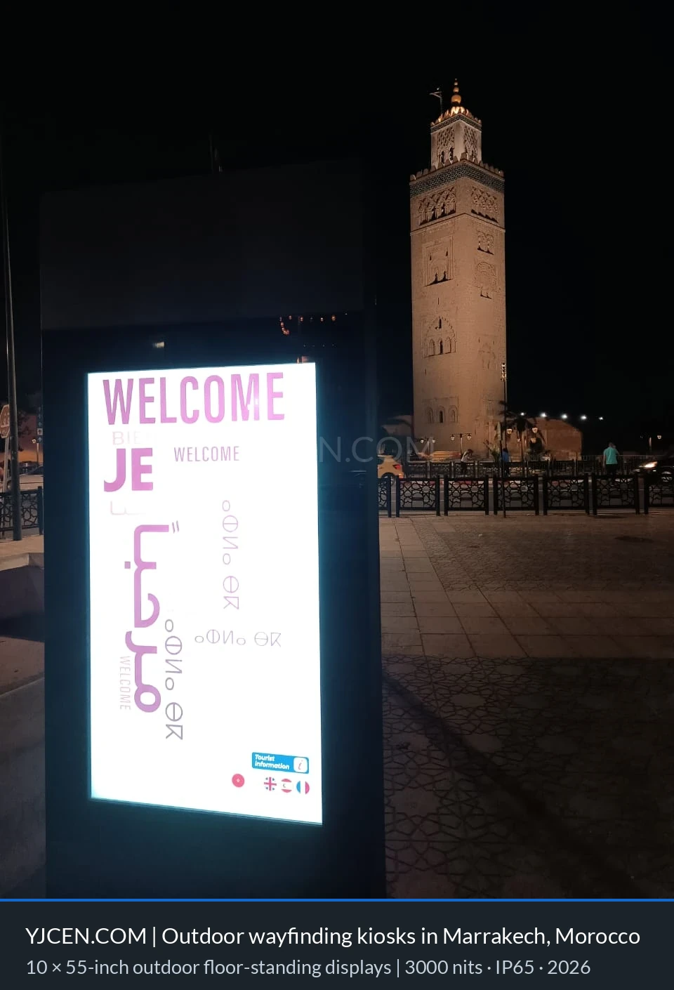 Nattvy av en utomhus turistinformationskiosk på 55 tum i Marrakech med belyst välkomstskärm och den upplysta Koutoubia-minareten bakom.