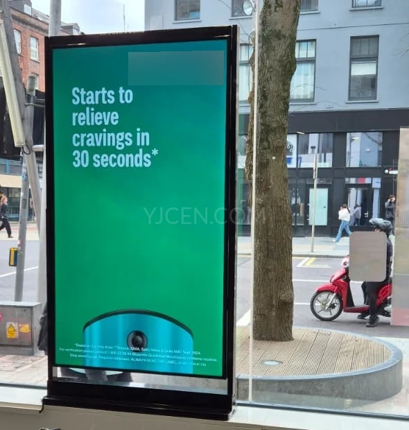 Display de vitrine vertical com conteúdo promocional verde ao lado de uma janela voltada para a rua.
