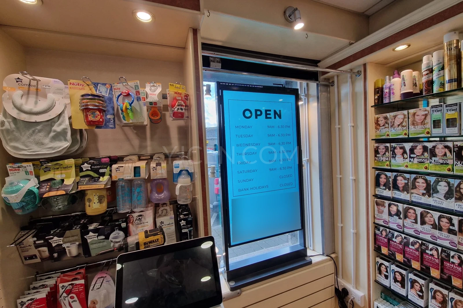 Display de vitrine vertical com horário de funcionamento sobre fundo azul entre prateleiras de uma loja.